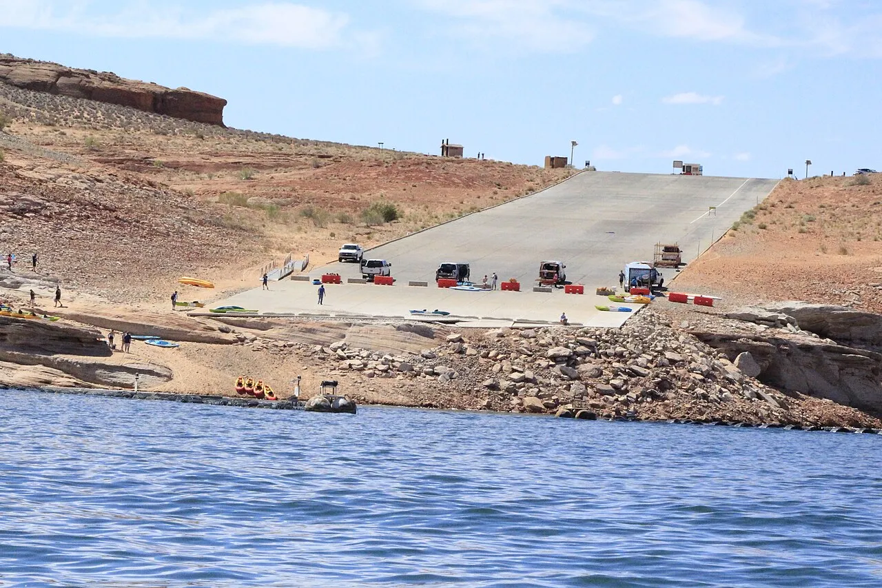 Trucks unloading kayaks on a barricaded launch ramp as visitors descend a rocky shoreline.